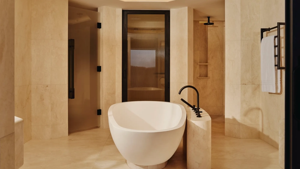 Freestanding oval bathtub in beige marble bathroom with rainfall shower and soft lighting.