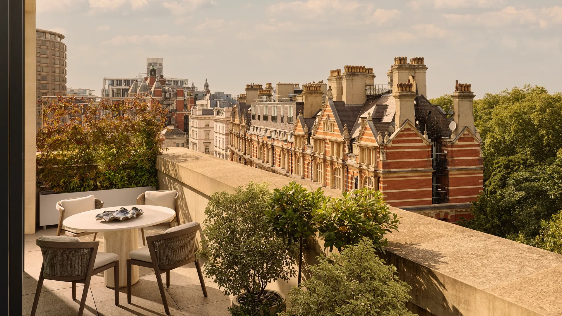 Outdoor terrace with round dining table and four chairs, bordered by potted greenery, overlooking historic red-brick buildings and Hyde Park.