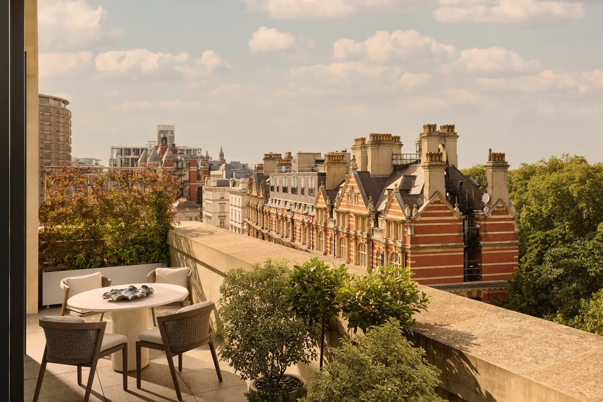 Outdoor terrace with round dining table and four chairs, bordered by potted greenery, overlooking historic red-brick buildings and Hyde Park.