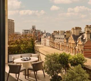 Outdoor terrace with round dining table and four chairs, bordered by potted greenery, overlooking historic red-brick buildings and Hyde Park.