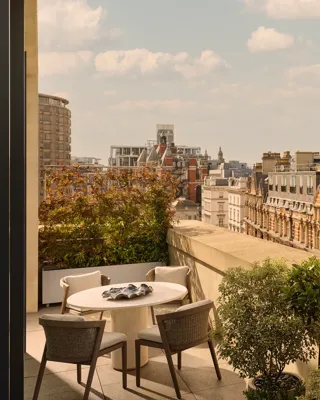 Outdoor terrace with round dining table and four chairs, bordered by potted greenery, overlooking historic red-brick buildings and Hyde Park.