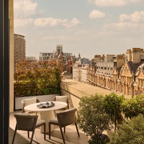Outdoor terrace with round dining table and four chairs, bordered by potted greenery, overlooking historic red-brick buildings and Hyde Park.