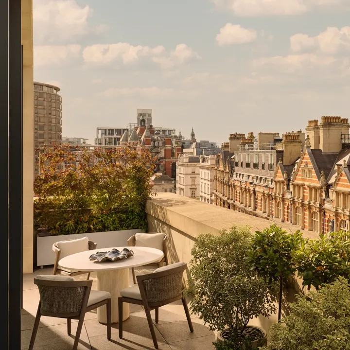 Outdoor terrace with round dining table and four chairs, bordered by potted greenery, overlooking historic red-brick buildings and Hyde Park.