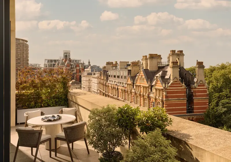 Outdoor terrace with round dining table and four chairs, bordered by potted greenery, overlooking historic red-brick buildings and Hyde Park.
