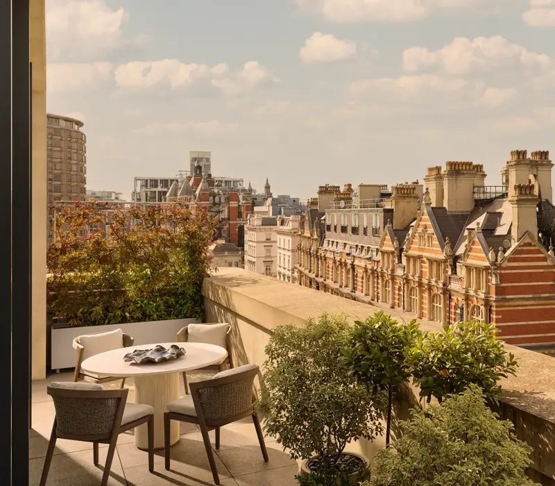 Outdoor terrace with round dining table and four chairs, bordered by potted greenery, overlooking historic red-brick buildings and Hyde Park.