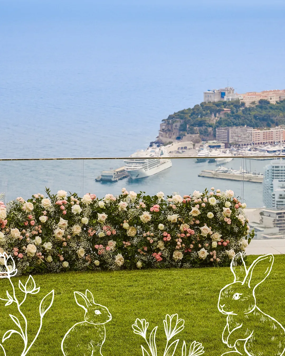 Terrace with floral hedge overlooking Monaco harbor, cruise ship and hillside buildings, with decorative rabbit illustrations on grass.