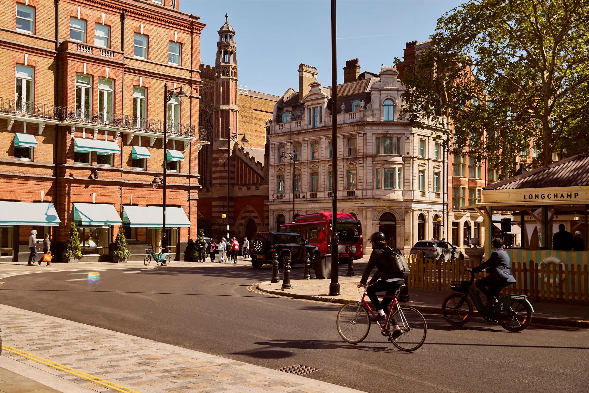 Cyclists and traffic move through a sunlit city square lined with historic buildings, shops, and mature trees.