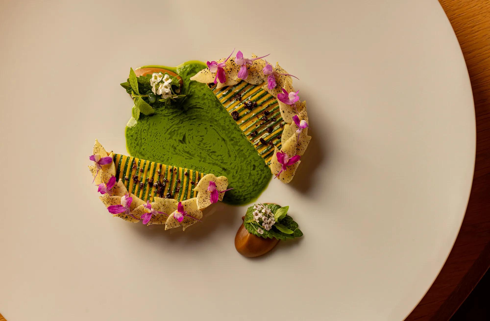 Artfully plated dish with green sauce, layered vegetable slices, and small edible flowers on a white plate.