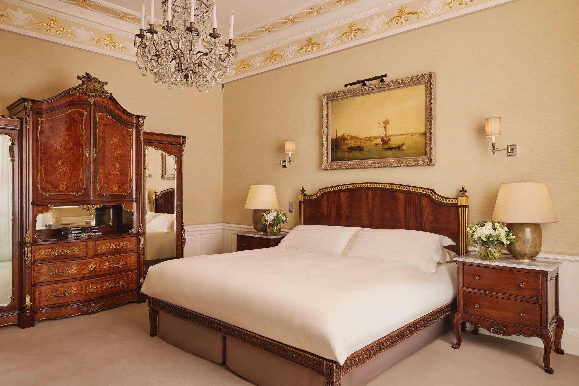 Elegant bedroom with carved wood bed, antique wardrobe, chandelier, and framed artwork above the headboard.