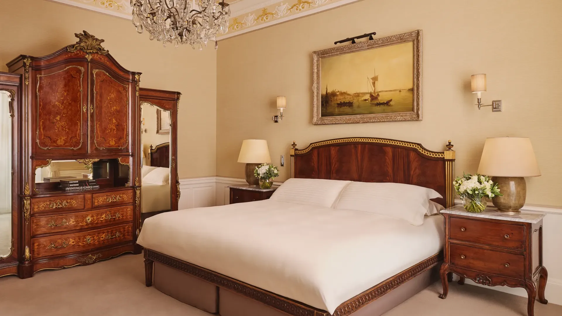 Elegant bedroom with carved wood bed, antique wardrobe, chandelier, and framed artwork above the headboard.