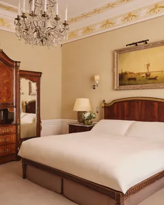 Elegant bedroom with carved wood bed, antique wardrobe, chandelier, and framed artwork above the headboard.