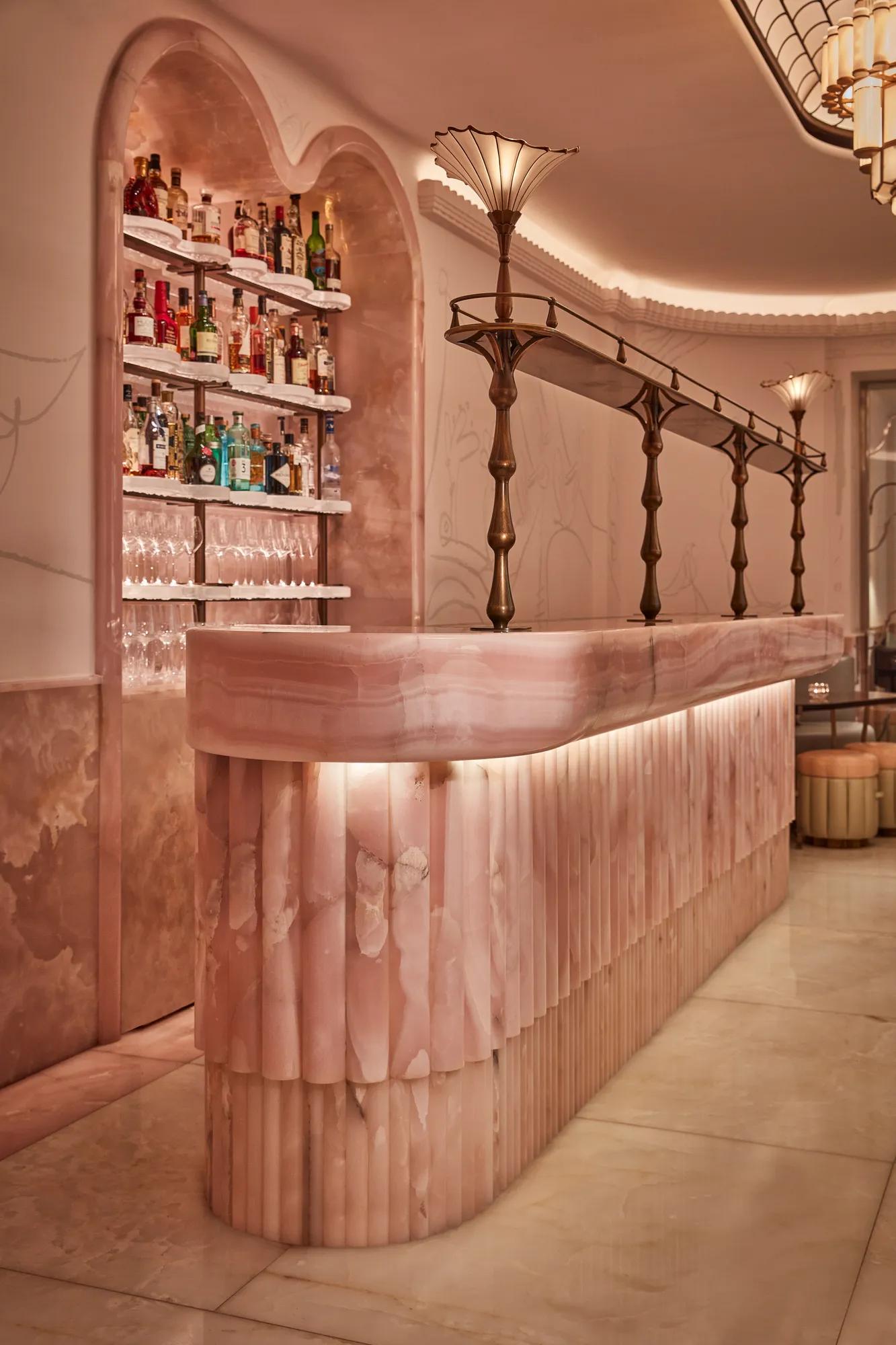 Art deco–inspired bar with a sculpted pink onyx counter, brass detailing, and soft ambient lighting. The backlit shelves display bottles of spirits and glassware, adding elegance to the intimate, pastel-toned space.