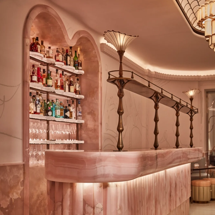 Art deco–inspired bar with a sculpted pink onyx counter, brass detailing, and soft ambient lighting. The backlit shelves display bottles of spirits and glassware, adding elegance to the intimate, pastel-toned space.