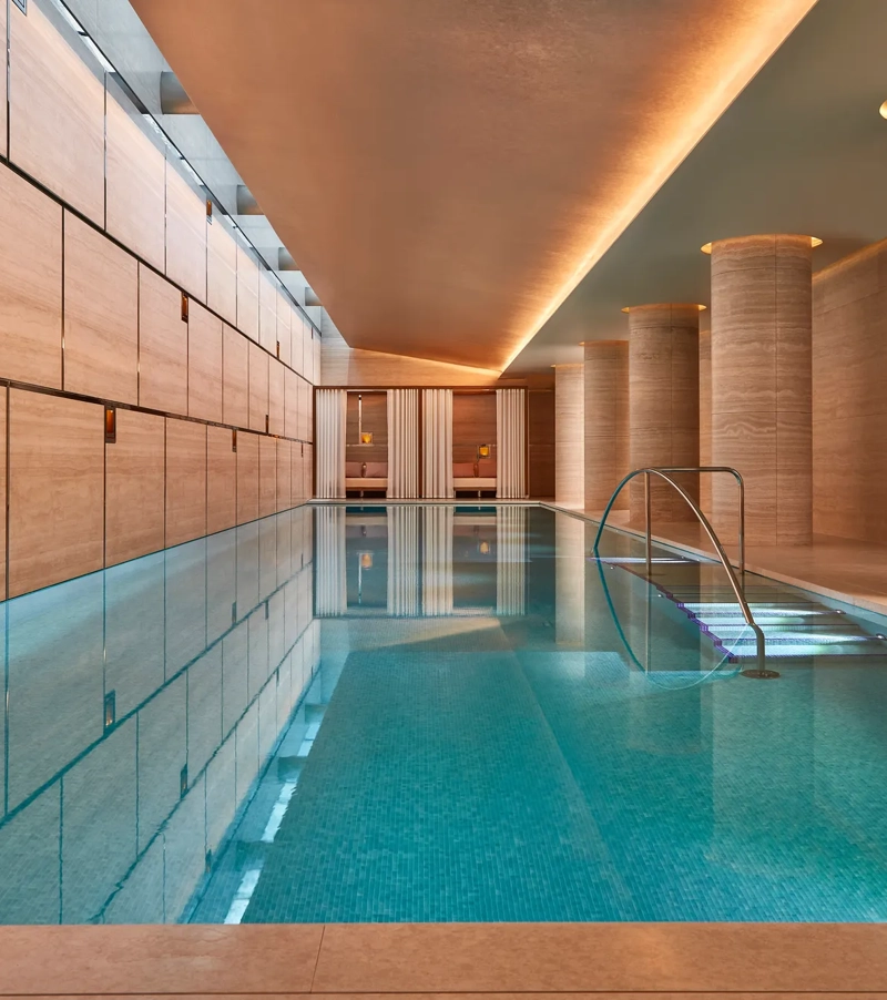 Indoor lap pool with travertine columns, soft ceiling lighting, and private curtained cabanas at the far end.