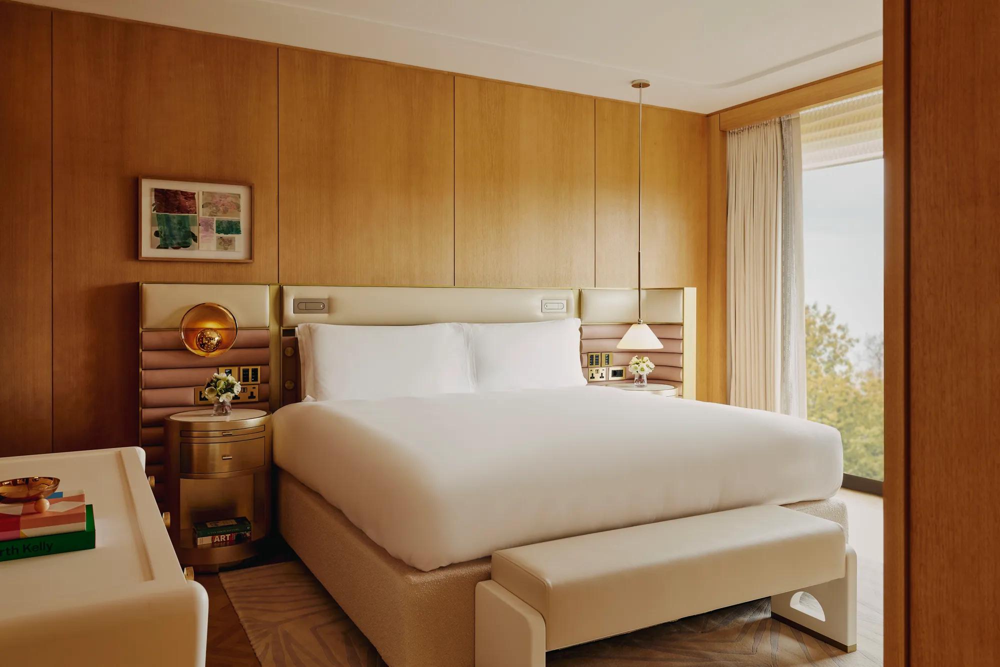 Hotel bedroom with king bed, upholstered headboard, wood-panelled walls, bedside lighting and large window with treetop views.