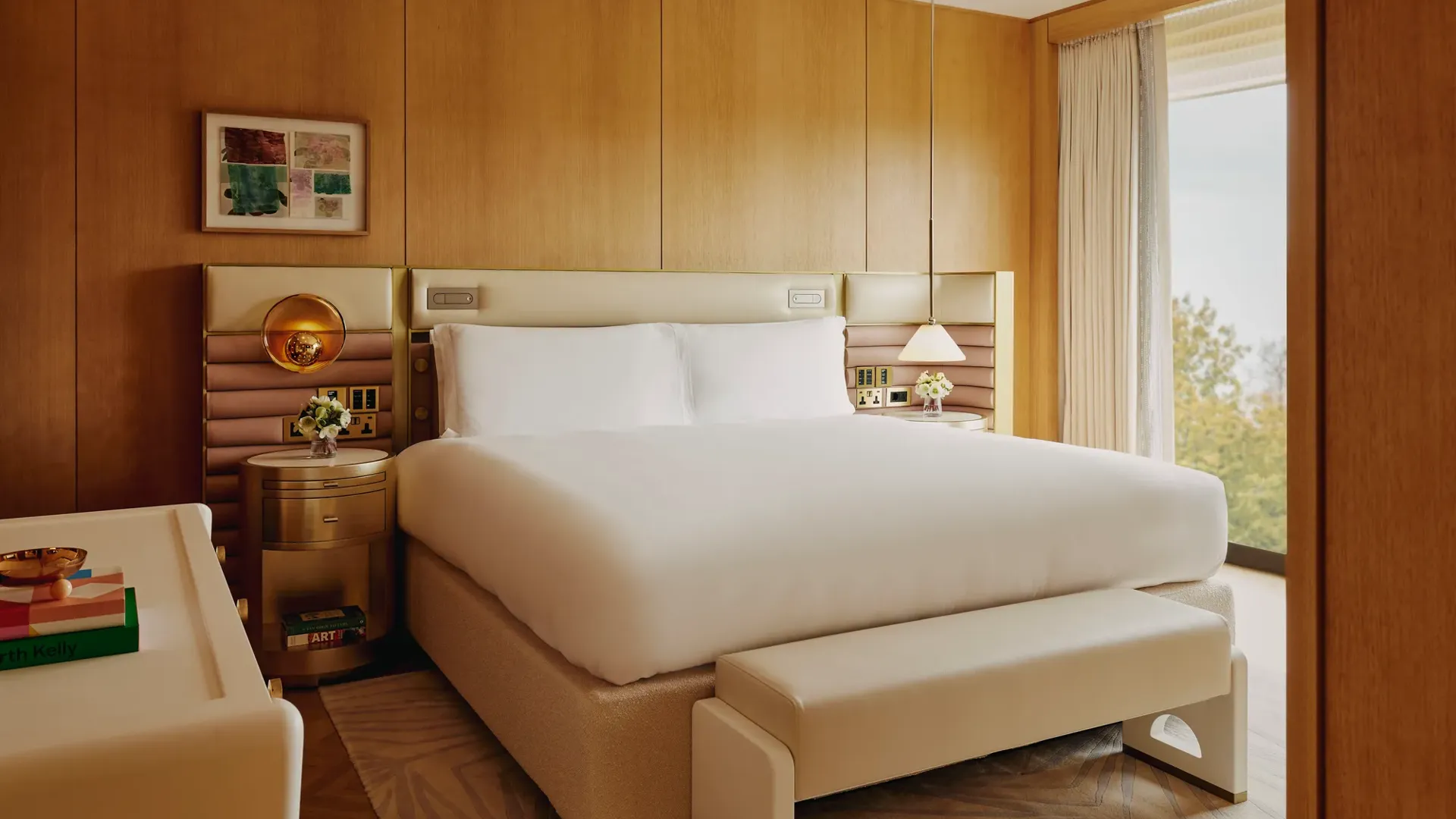 Hotel bedroom with king bed, upholstered headboard, wood-panelled walls, bedside lighting and large window with treetop views.