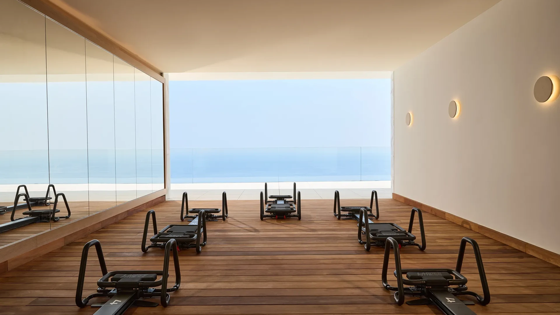 Modern fitness studio with reformer machines on wooden flooring, mirrored wall, and open view of the sea through a glass-fronted terrace.