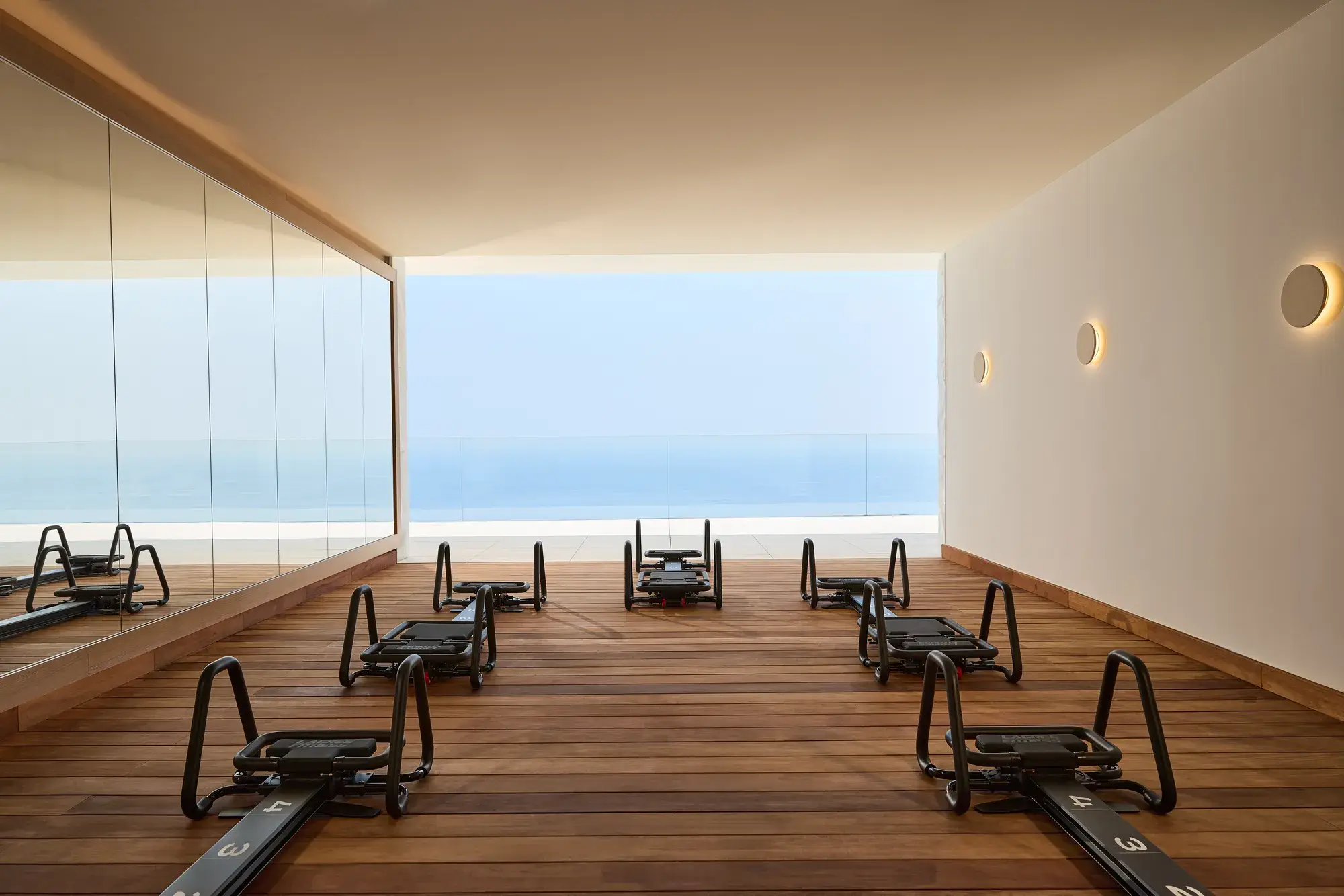 Modern fitness studio with reformer machines on wooden flooring, mirrored wall, and open view of the sea through a glass-fronted terrace.