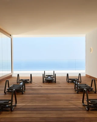 Modern fitness studio with reformer machines on wooden flooring, mirrored wall, and open view of the sea through a glass-fronted terrace.
