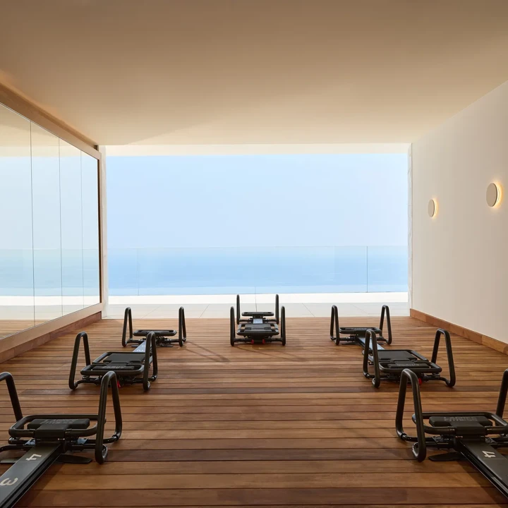 Modern fitness studio with reformer machines on wooden flooring, mirrored wall, and open view of the sea through a glass-fronted terrace.