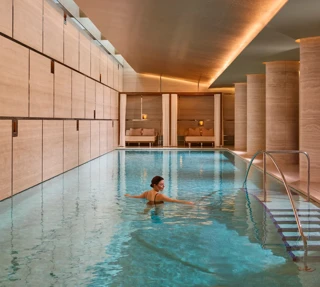 Guest swimming in a serene indoor spa pool with stone columns, warm lighting and steps with handrail.