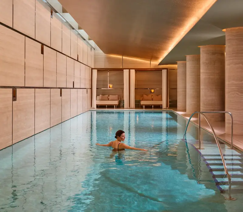 Guest swimming in a serene indoor spa pool with stone columns, warm lighting and steps with handrail.