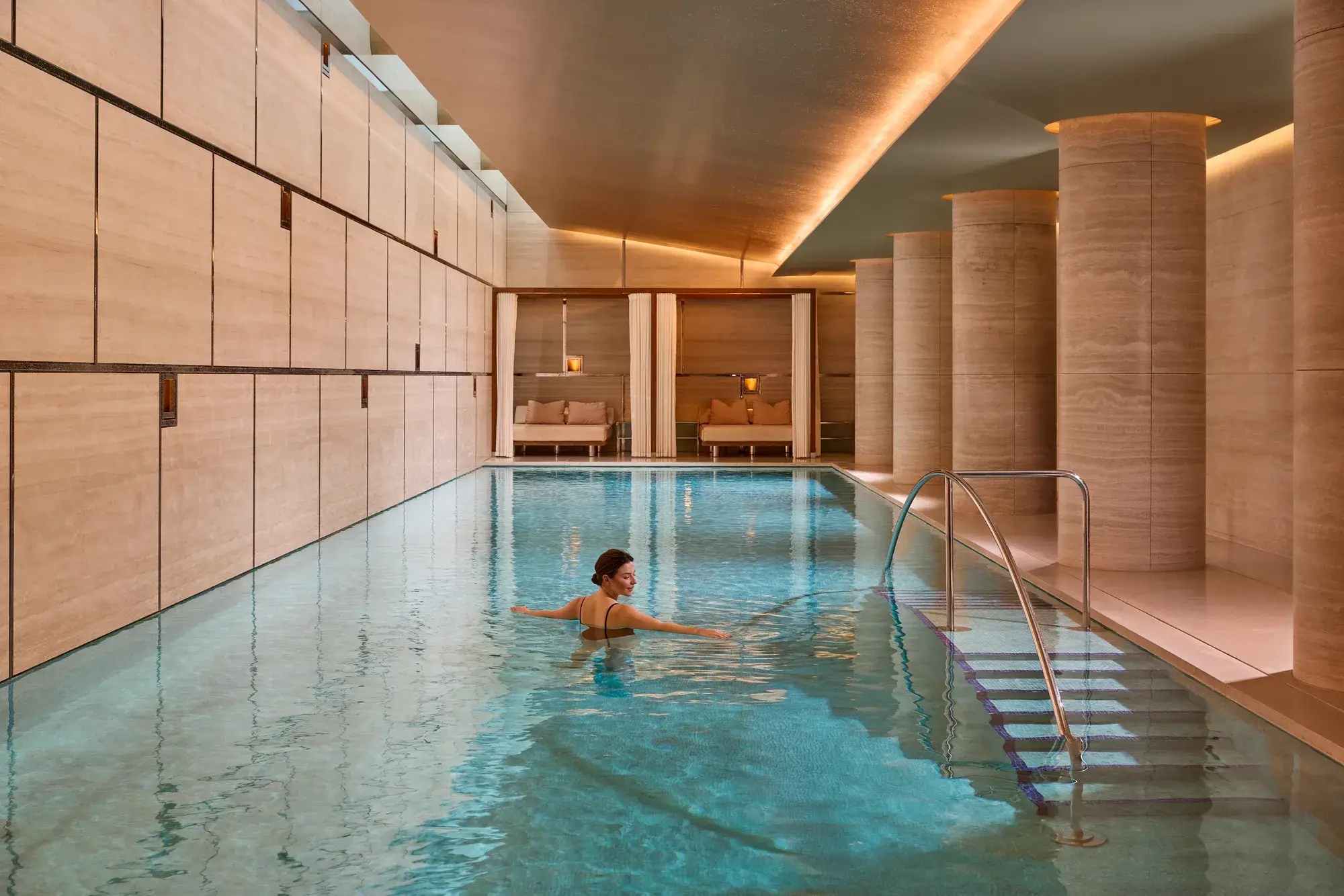 Guest swimming in a serene indoor spa pool with stone columns, warm lighting and steps with handrail.