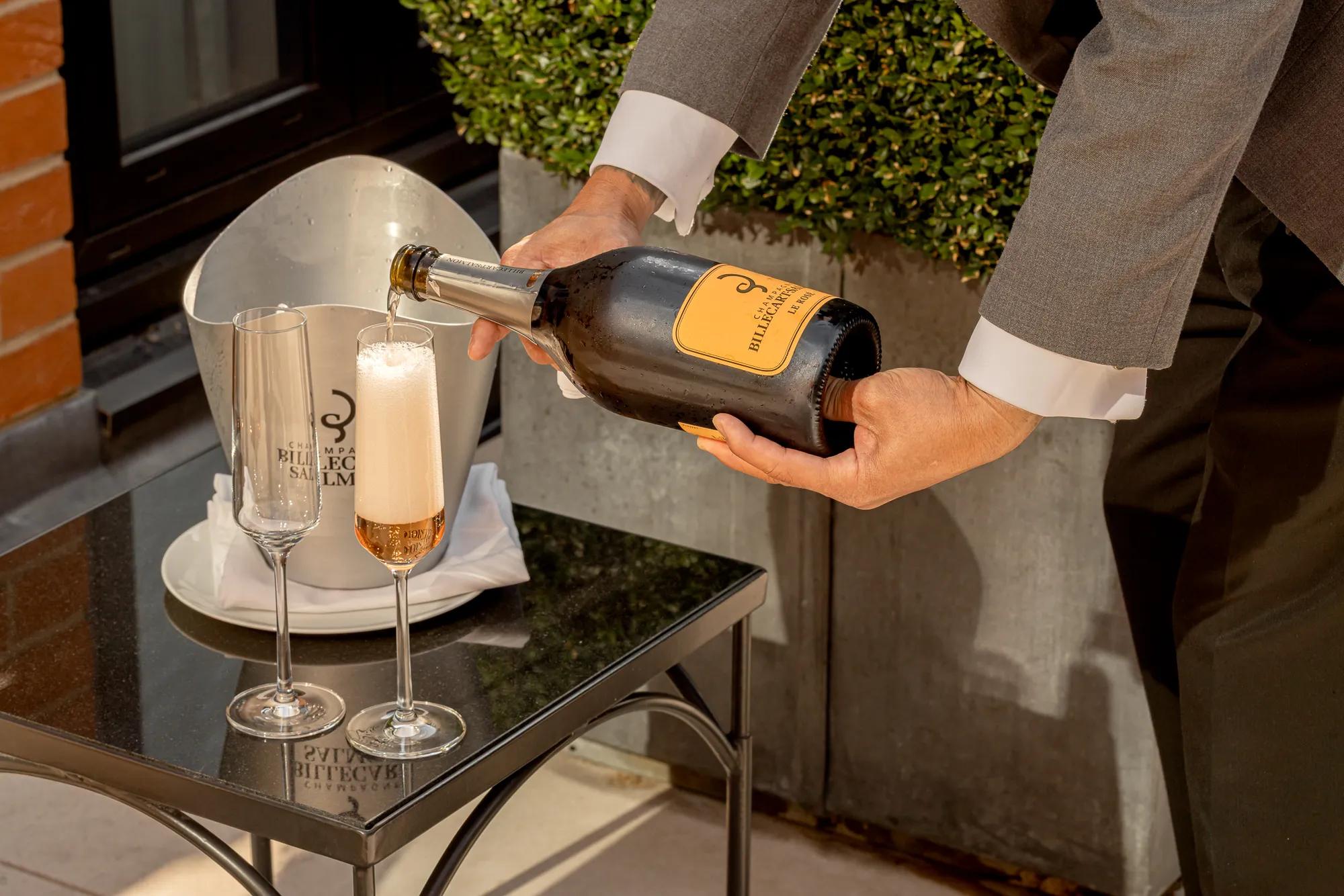Person pouring sparkling wine into a flute beside another empty glass, with ice bucket on small outdoor table and greenery in background.