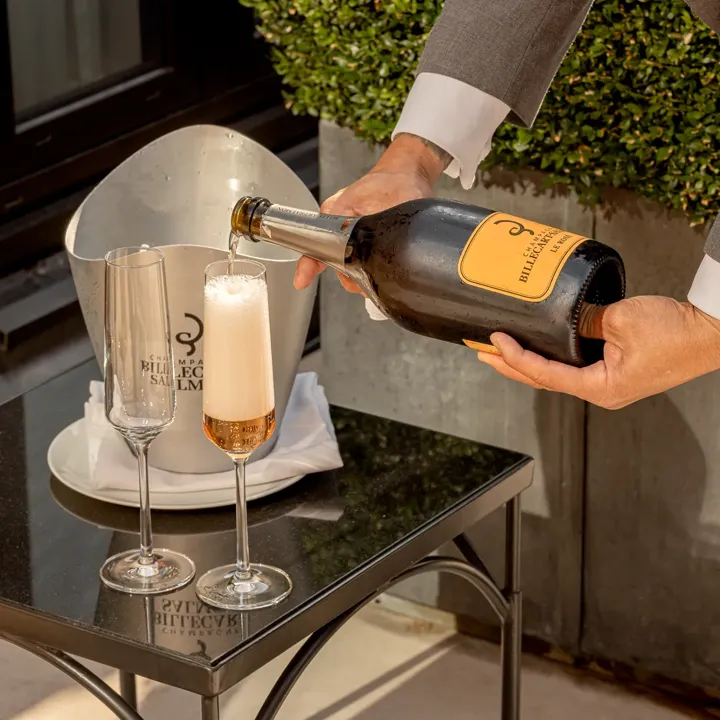 Person pouring sparkling wine into a flute beside another empty glass, with ice bucket on small outdoor table and greenery in background.