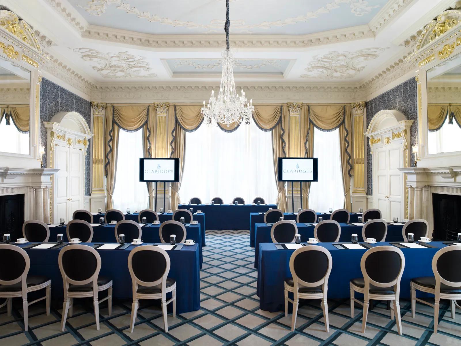 A grand meeting room at Claridge’s arranged in a conference setup with blue tablecloths, elegant chairs, and two presentation screens, featuring ornate gold detailing, chandeliers, and tall windows draped with gold curtains.