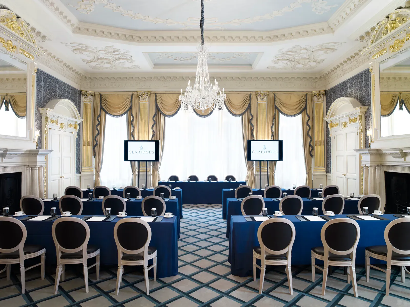 A grand meeting room at Claridge’s arranged in a conference setup with blue tablecloths, elegant chairs, and two presentation screens, featuring ornate gold detailing, chandeliers, and tall windows draped with gold curtains.