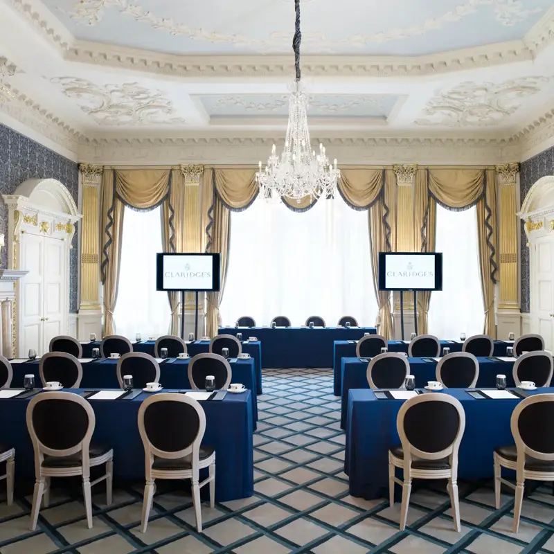 A grand meeting room at Claridge’s arranged in a conference setup with blue tablecloths, elegant chairs, and two presentation screens, featuring ornate gold detailing, chandeliers, and tall windows draped with gold curtains.