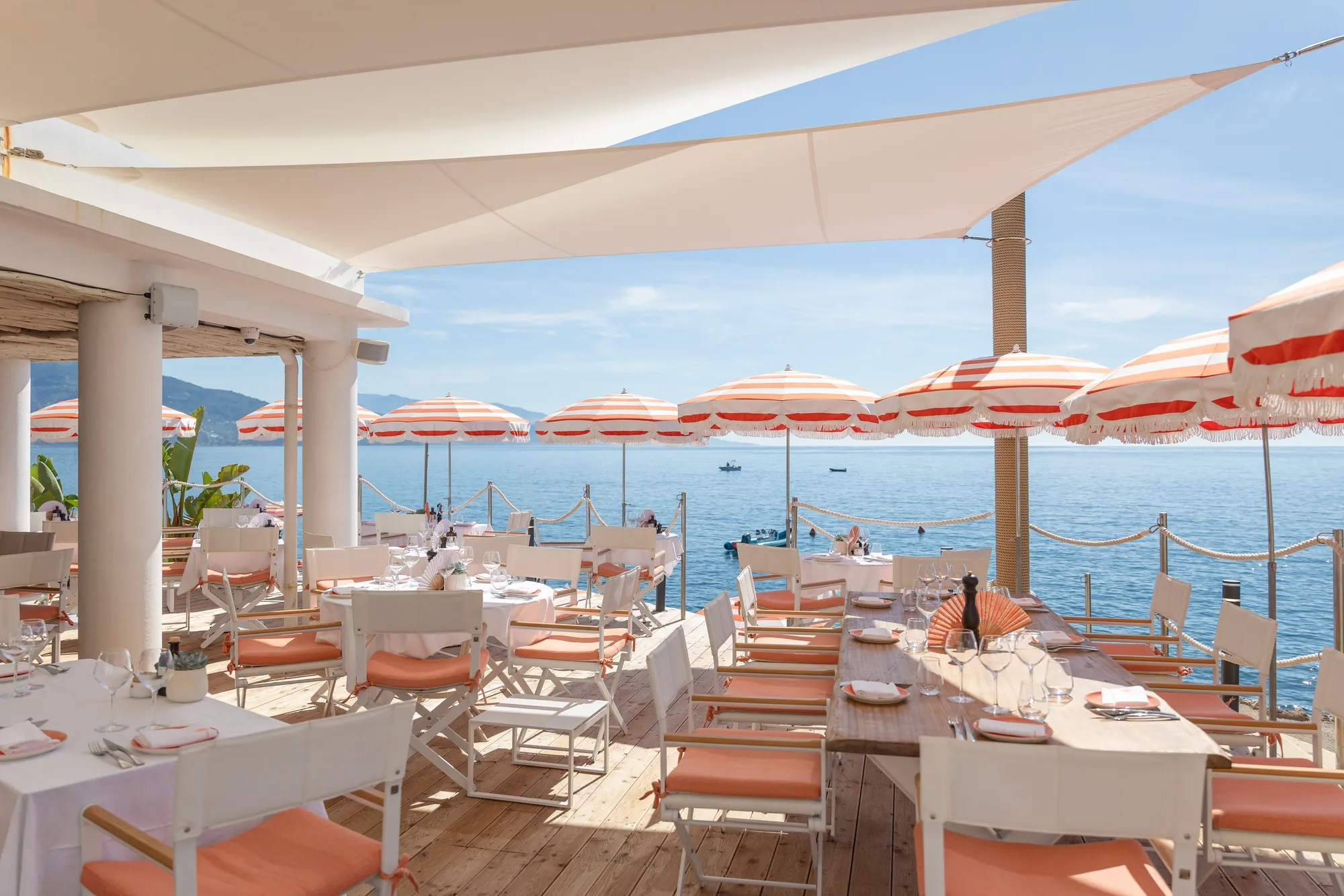Terrasse de restaurant en bord de mer avec tables blanches, chaises corail et parasols rayés face à la Méditerranée.