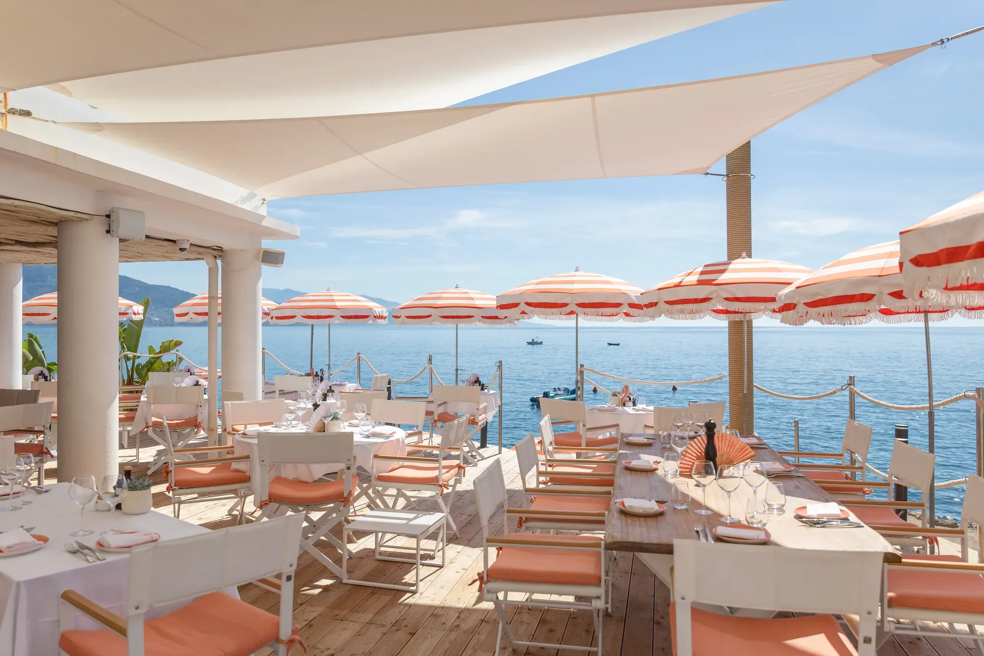 Terrasse de restaurant en bord de mer avec tables blanches, chaises corail et parasols rayés face à la Méditerranée.