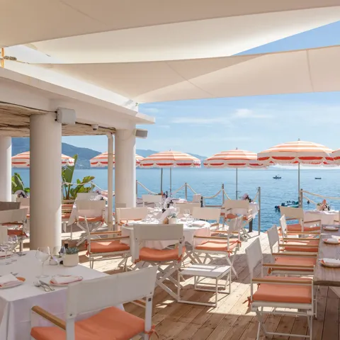 Terrasse de restaurant en bord de mer avec tables blanches, chaises corail et parasols rayés face à la Méditerranée.