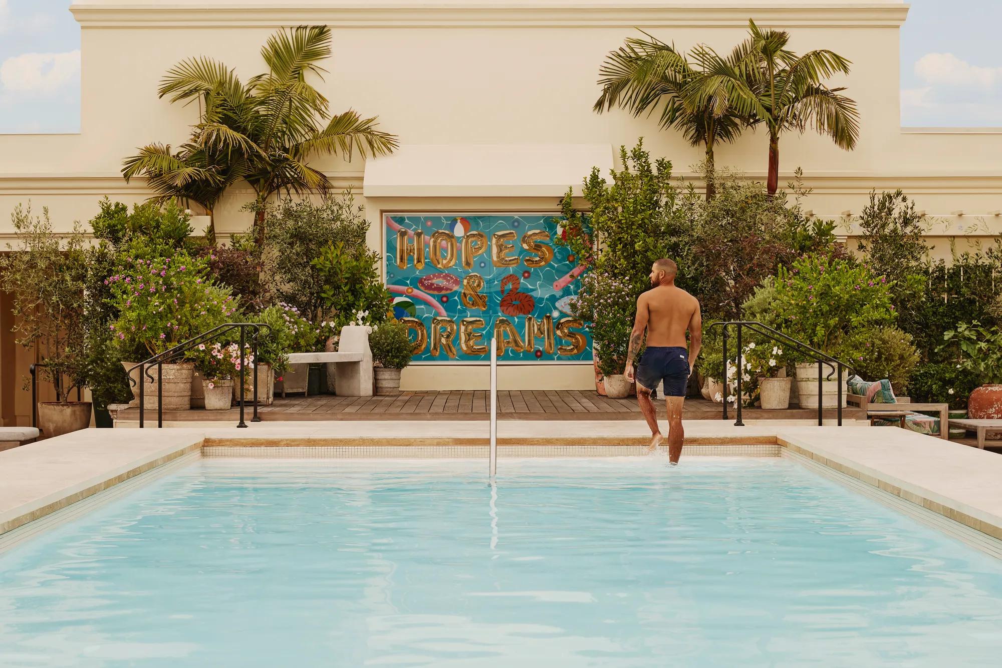 Guest stepping from The Maybourne Beverly Hills rooftop pool toward the “Hopes & Dreams” mural, palms and planters behind.