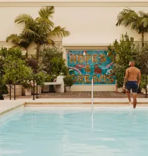 Guest stepping from The Maybourne Beverly Hills rooftop pool toward the “Hopes & Dreams” mural, palms and planters behind.