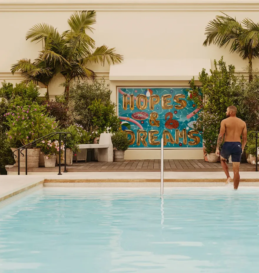 Guest stepping from The Maybourne Beverly Hills rooftop pool toward the “Hopes & Dreams” mural, palms and planters behind.