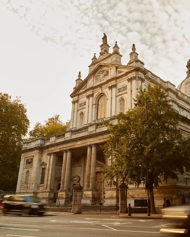 Large stone church with classical columns and dome, framed by trees and London taxis passing