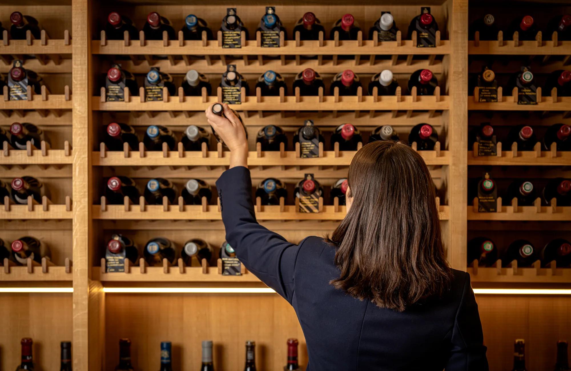 A wine specialist selects a bottle from a wall of wooden wine racks filled with bottles in a temperature-controlled wine cellar.