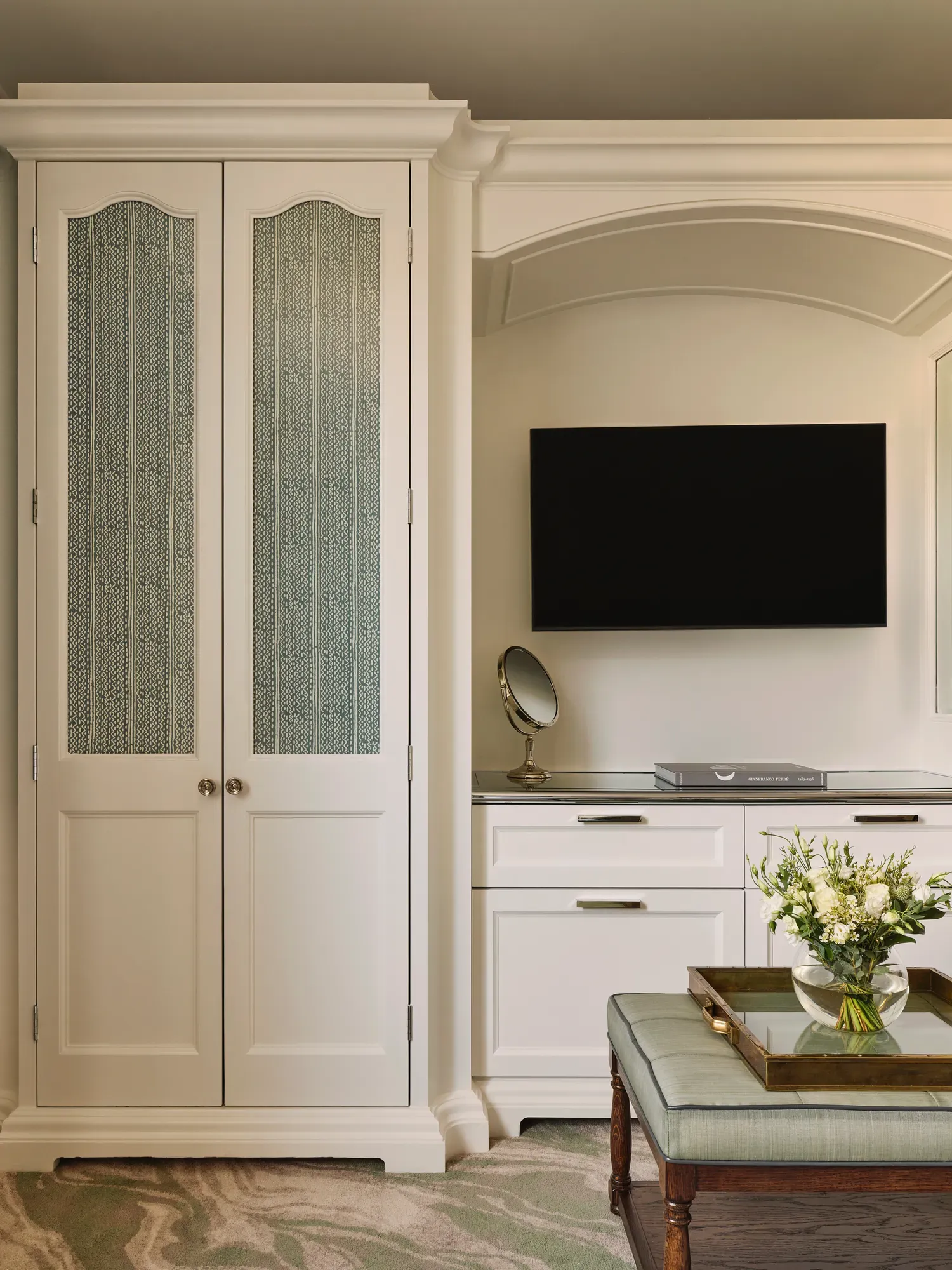 A built-in white wardrobe and dresser with a wall-mounted TV and a small ottoman topped with flowers on a tray.