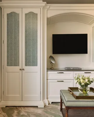 A built-in white wardrobe and dresser with a wall-mounted TV and a small ottoman topped with flowers on a tray.