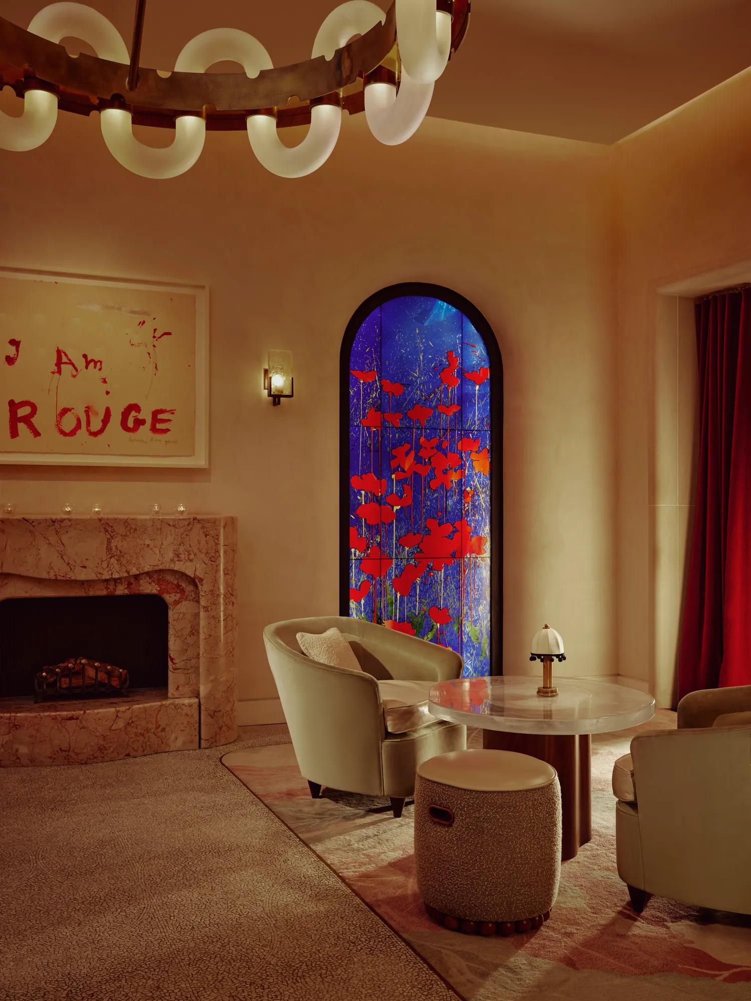 Elegant lounge with marble fireplace, sculptural lighting, plush seating and vivid stained-glass window featuring red floral motifs.