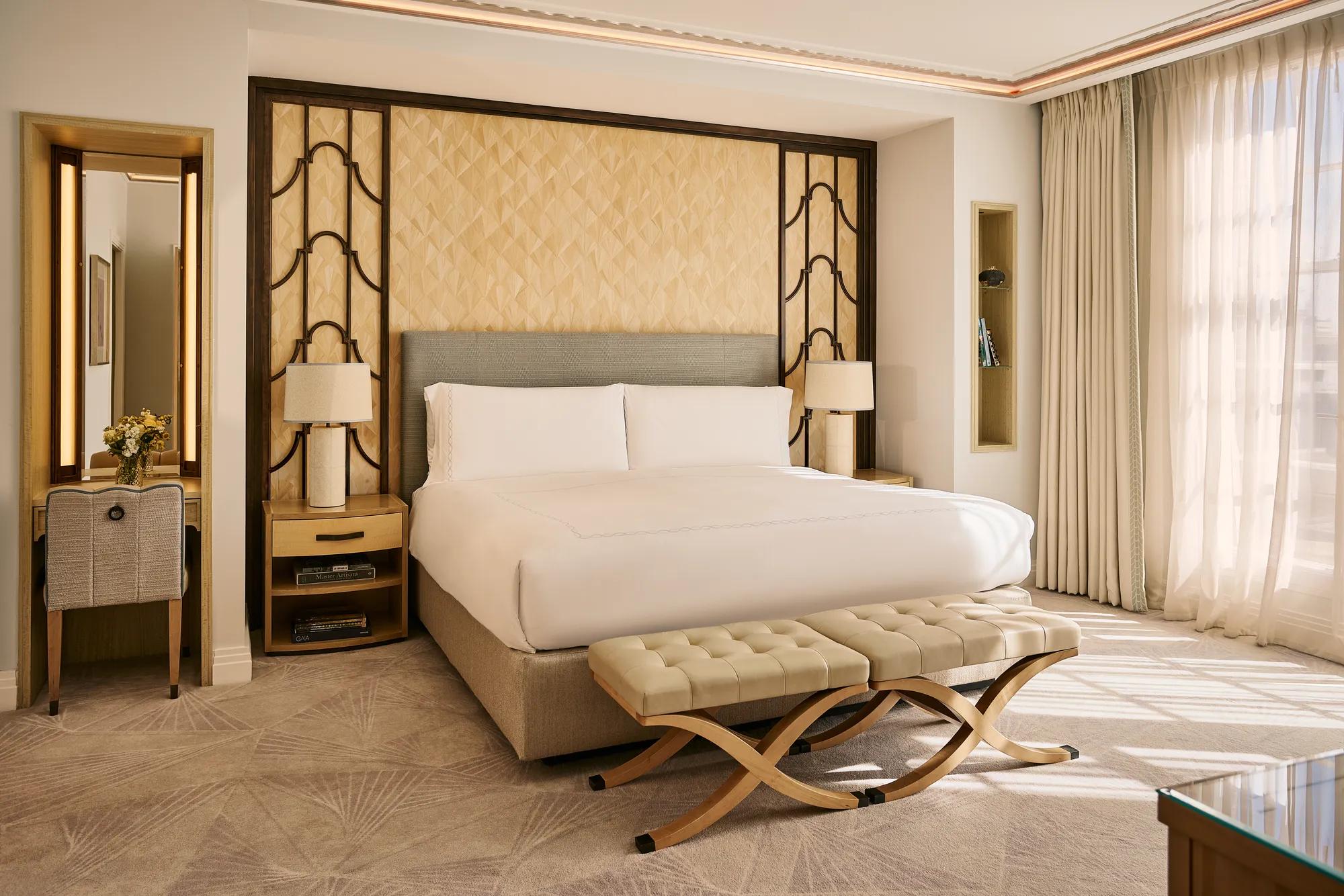 Bright bedroom with king bed, upholstered headboard, bedside tables with lamps, cushioned bench, and floor-to-ceiling window with curtains.