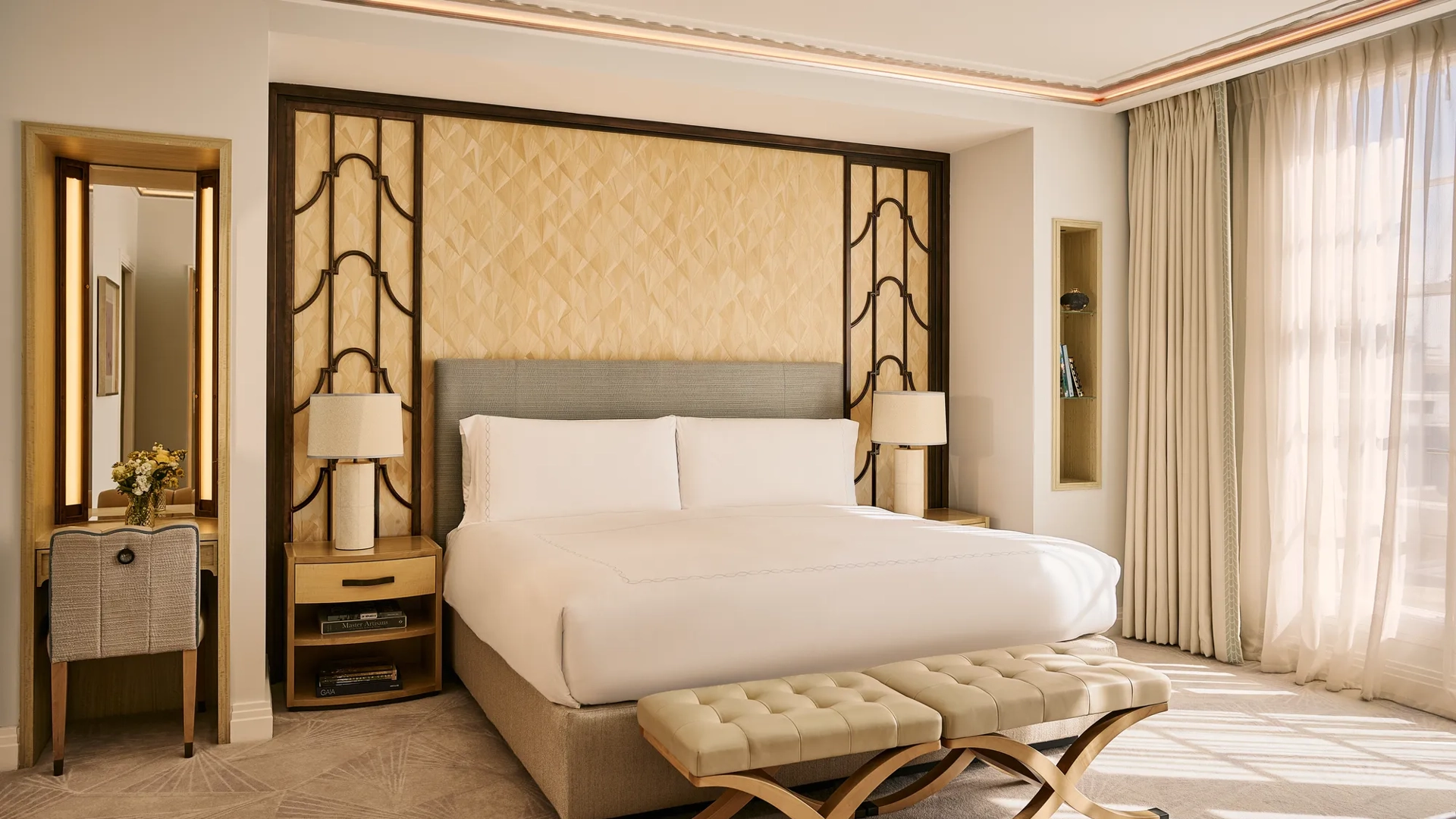 Bright bedroom with king bed, upholstered headboard, bedside tables with lamps, cushioned bench, and floor-to-ceiling window with curtains.