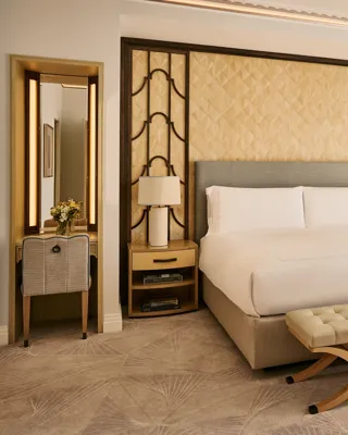 Bright bedroom with king bed, upholstered headboard, bedside tables with lamps, cushioned bench, and floor-to-ceiling window with curtains.