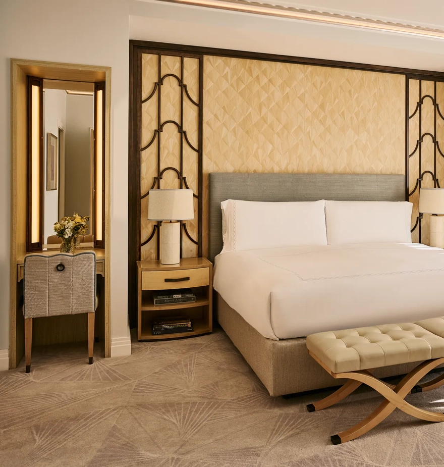 Bright bedroom with king bed, upholstered headboard, bedside tables with lamps, cushioned bench, and floor-to-ceiling window with curtains.