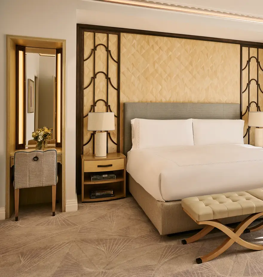 Bright bedroom with king bed, upholstered headboard, bedside tables with lamps, cushioned bench, and floor-to-ceiling window with curtains.