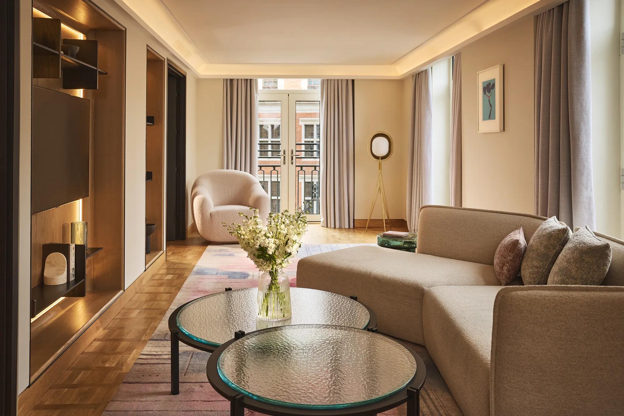 Bright hotel living room with sofa, armchair, round glass tables, and French doors opening to a balcony.