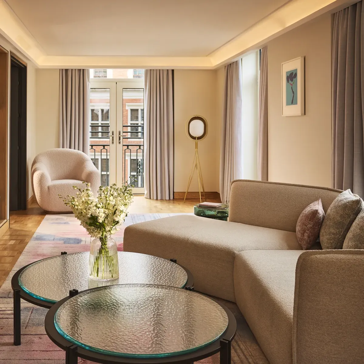 Bright hotel living room with sofa, armchair, round glass tables, and French doors opening to a balcony.